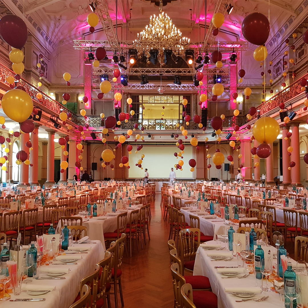 Festlich geschmückter Festsaal mit einer Tafel-Bestuhlung. Im Festsaal sind viele gelbe und rote Luftballons.