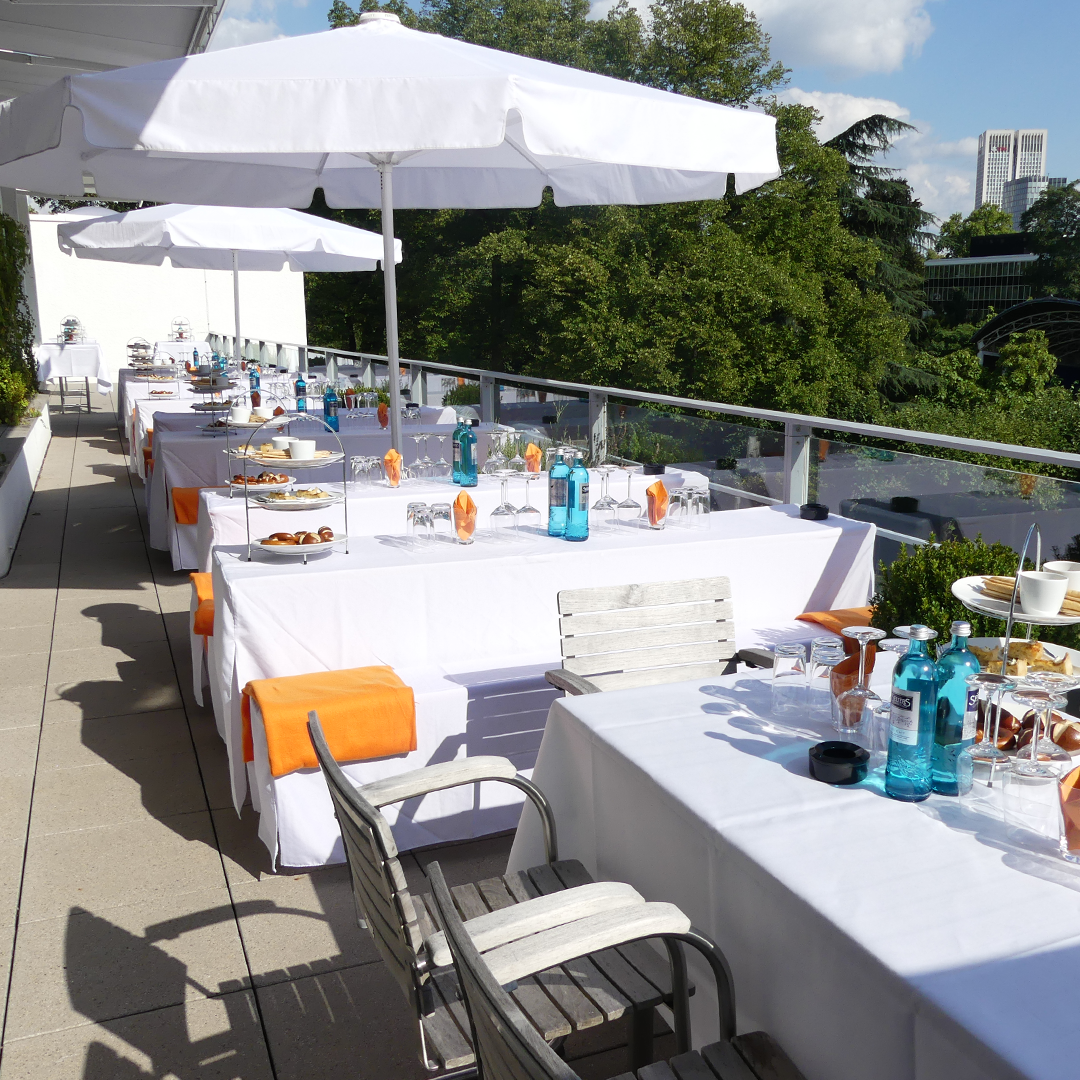 Terrasse mit festlich gedeckten Tischen. Die Tische sind elegant mit Tischdecken, Besteck, Gläsern und kleinen Mahlzeiten vorbereitet.