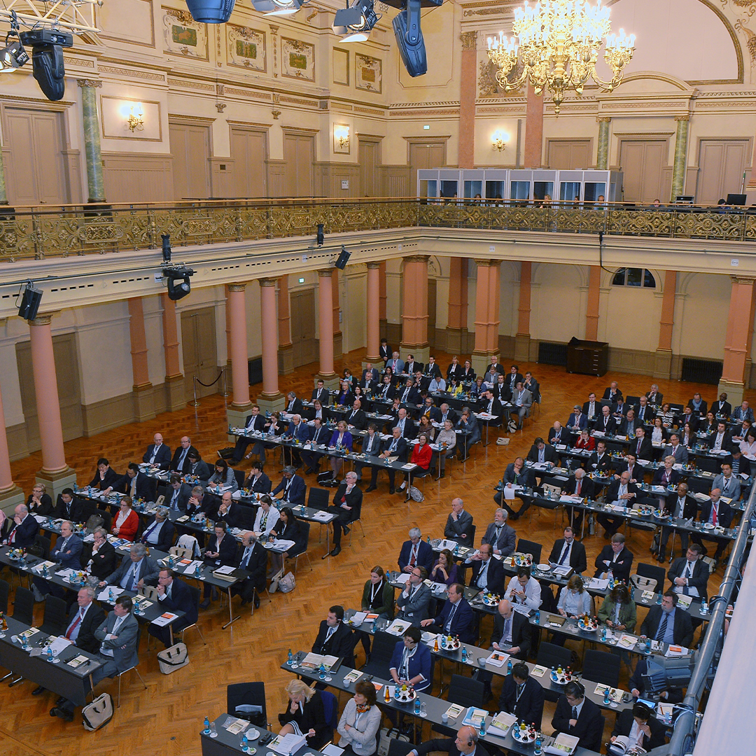 Parlamentarische Bestuhlung des Festsaals während einer Tagung. Mehrere Personen sitzen auf ihren Stühlen und verfolgen aufmerksam die Veranstaltung.