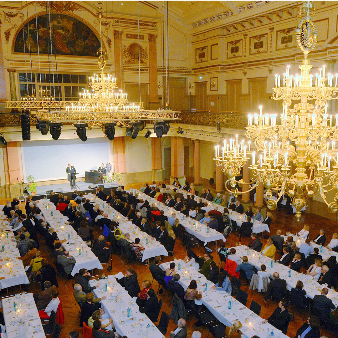 Mehrere Personen sitzen an langen Tafeln im Festsaal und blicken zur Bühne, auf der ein musikalisches Programm stattfindet.