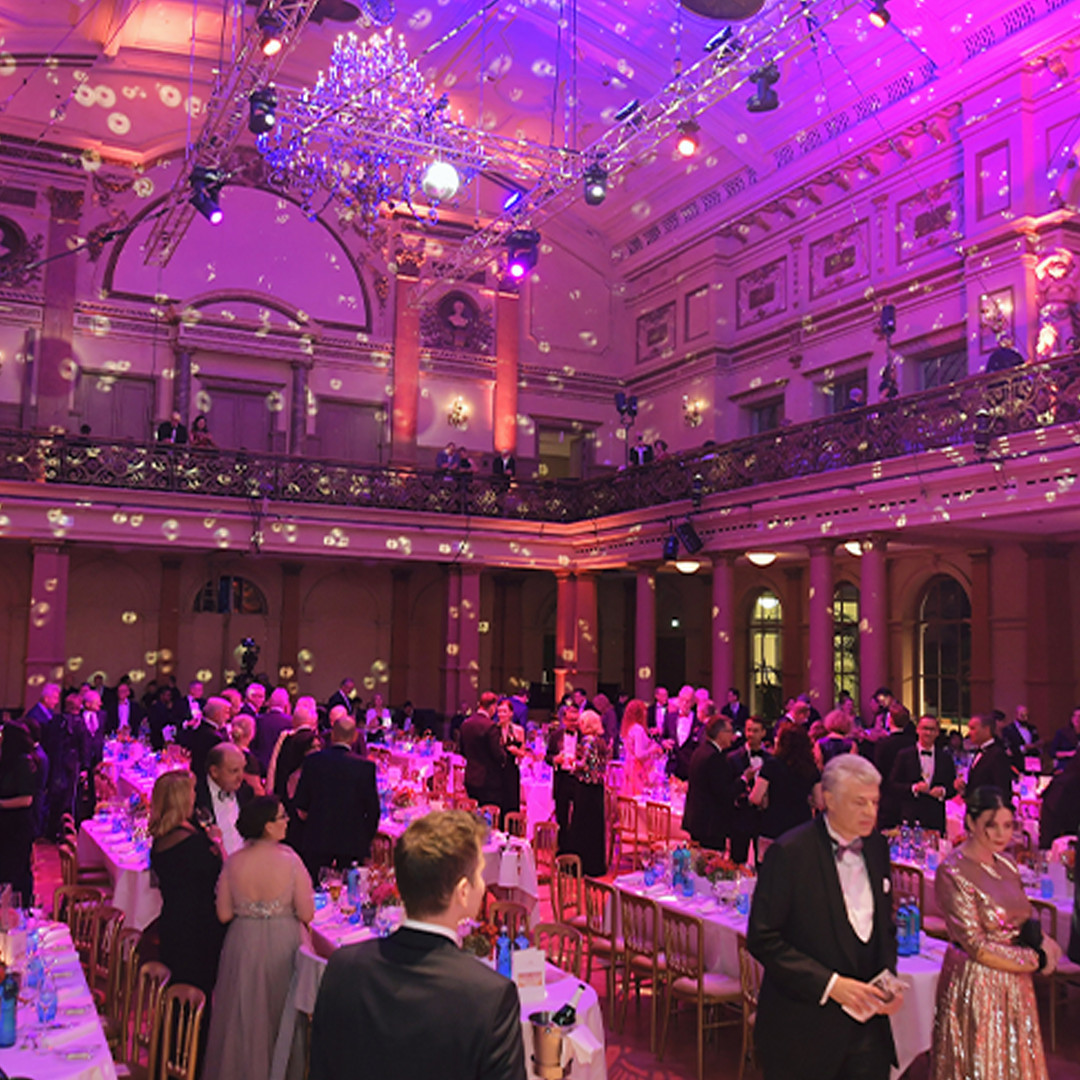 Festlich geschmückter Festsaal mit mehreren Personen in eleganten Outfits. Der Raum ist aufwendig dekoriert, und die Gäste genießen die feierliche Atmosphäre in ihren schicken Kleidungsstücken.