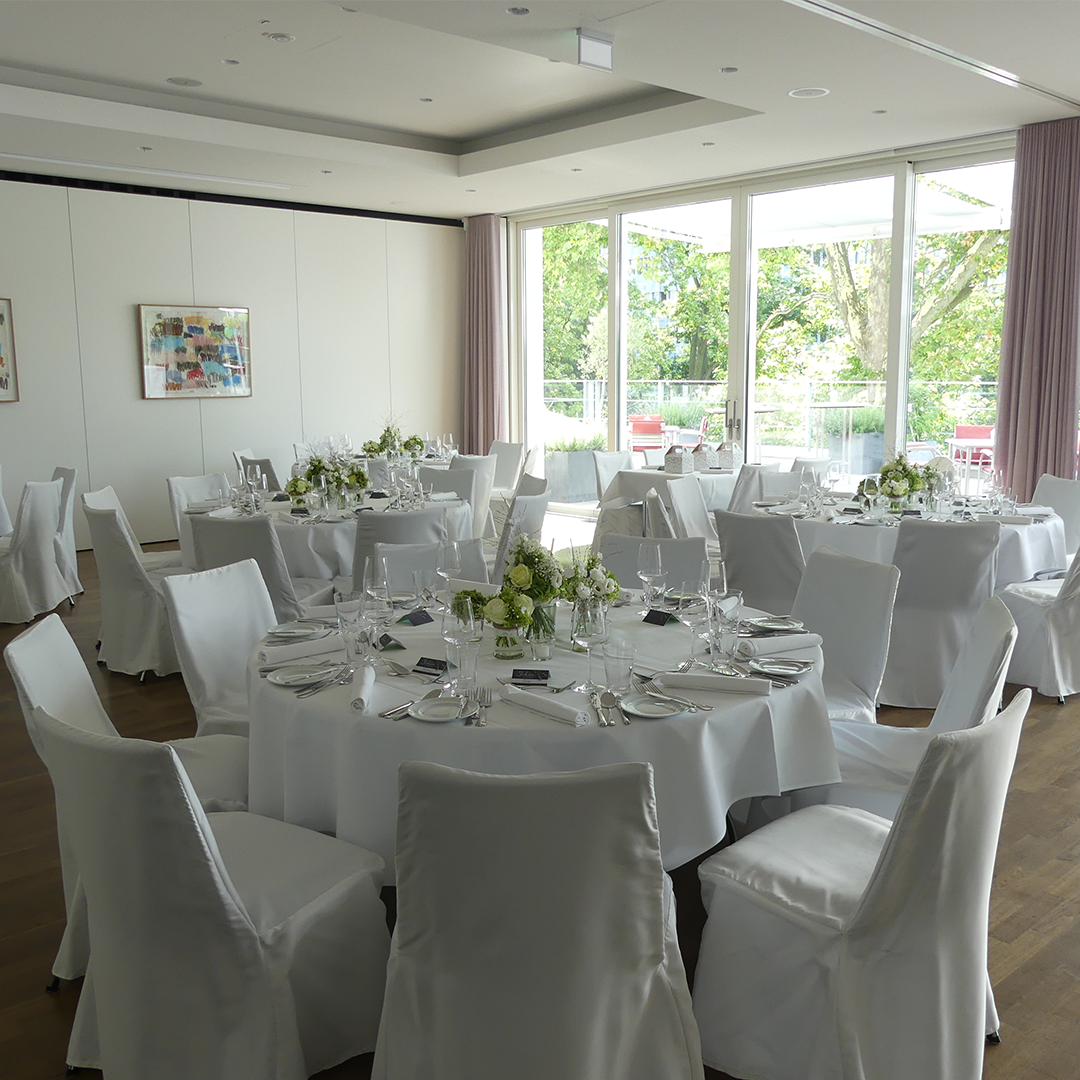 Text: Festlich gedeckter Raum „Orchidee“ mit weißer Tischdecke und elegantem Besteck. Der Raum bietet einen Blick auf die Terrasse, die durch große Fenster oder Türen sichtbar ist. Die Tischdekoration trägt zur eleganten Atmosphäre des Raums bei.
